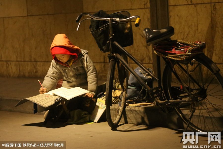 鄭州中原路京廣鐵路大橋下涵洞的馬路牙子邊，一位戴著紅色辣椒帽的小男孩趴在一個(gè)小馬扎上，上面鋪著一張車(chē)墊，在微弱的燈光下寫(xiě)作業(yè)